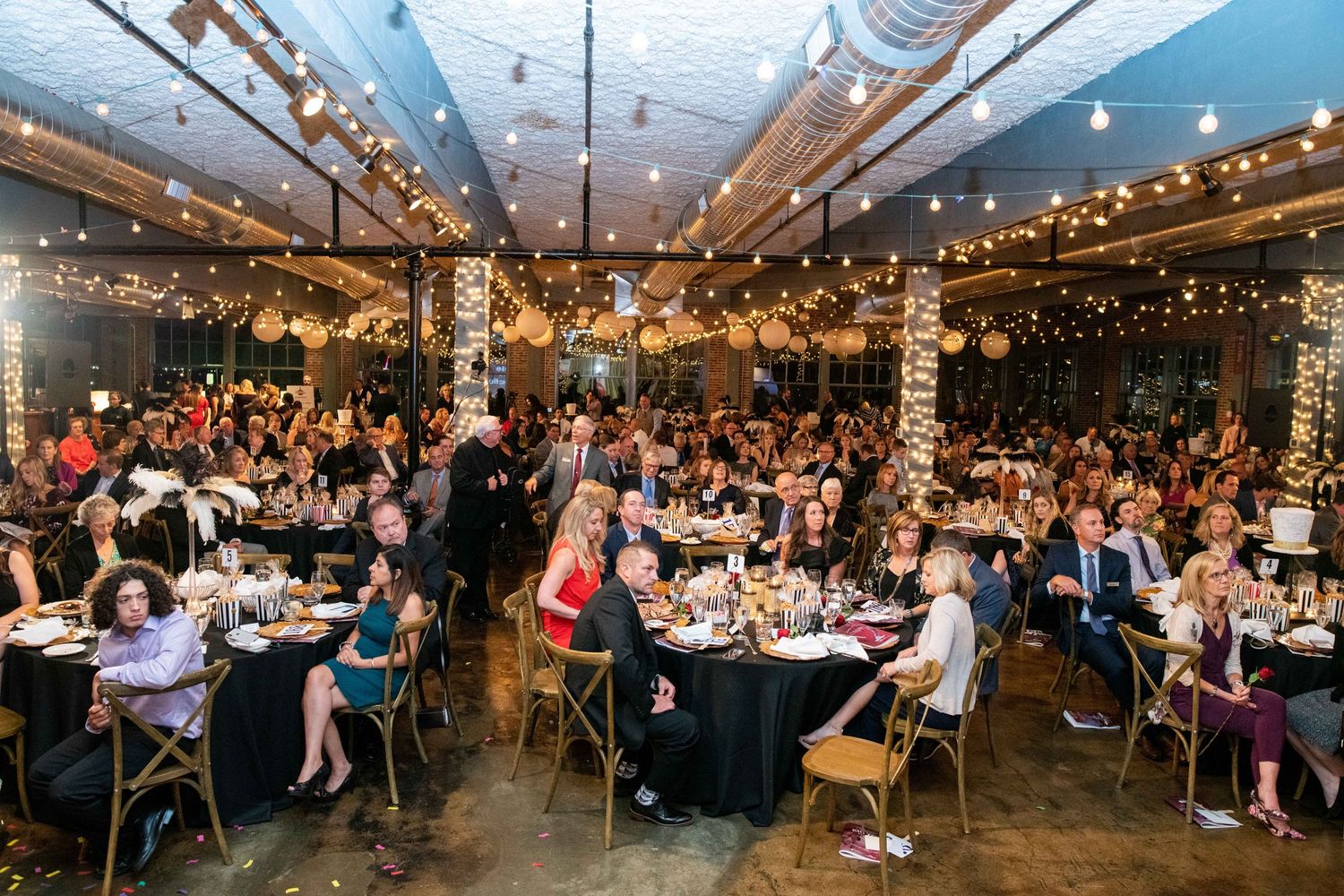 Large gathering in industrial venue with exposed beams and string lights during formal dinner event.
