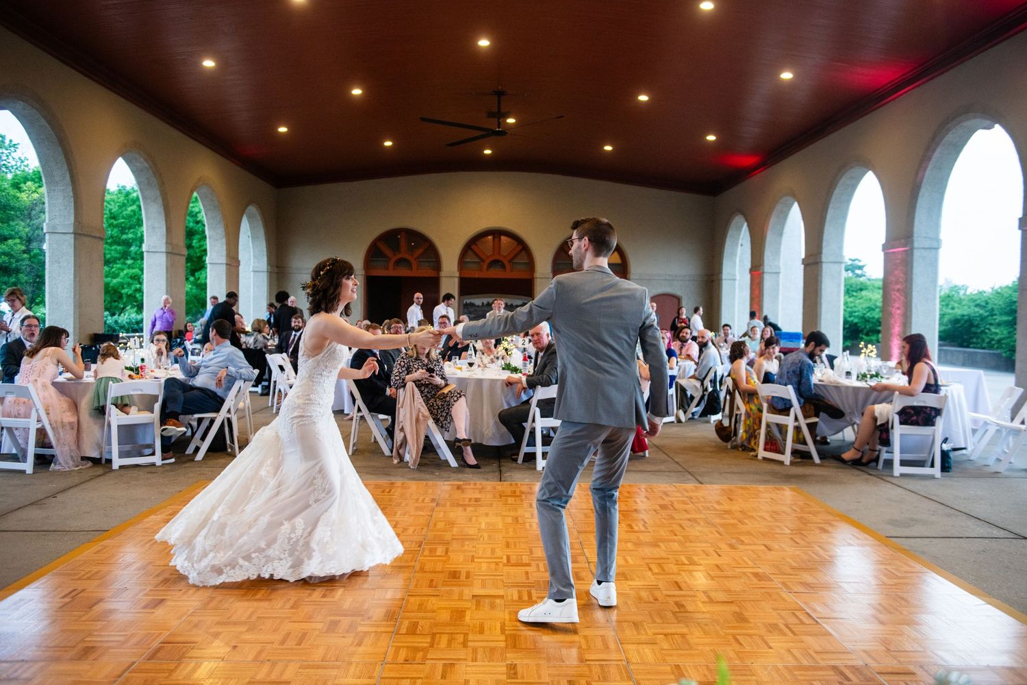 A couple performing their first dance in a reception hall with arched windows and watching guests.