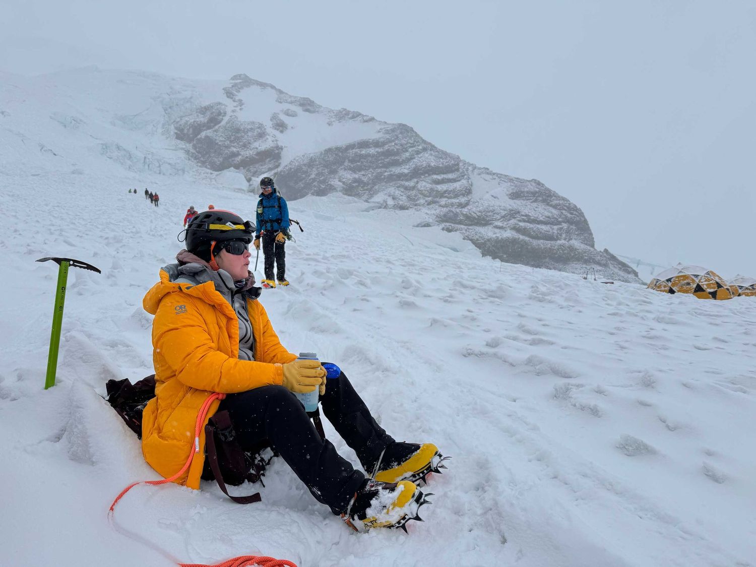Mountaineer in yellow jacket resting on snowy alpine slope with ice axe and crampons during a winter climb.