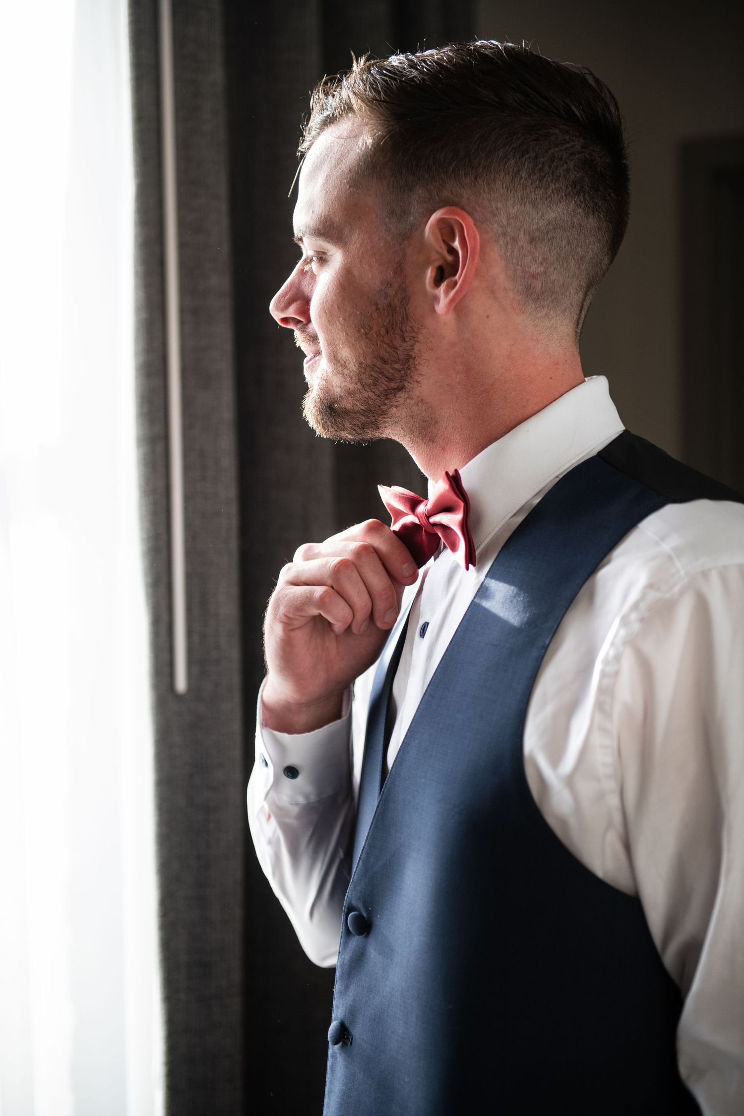 Side profile of groom in formal attire adjusting red bow tie by window.