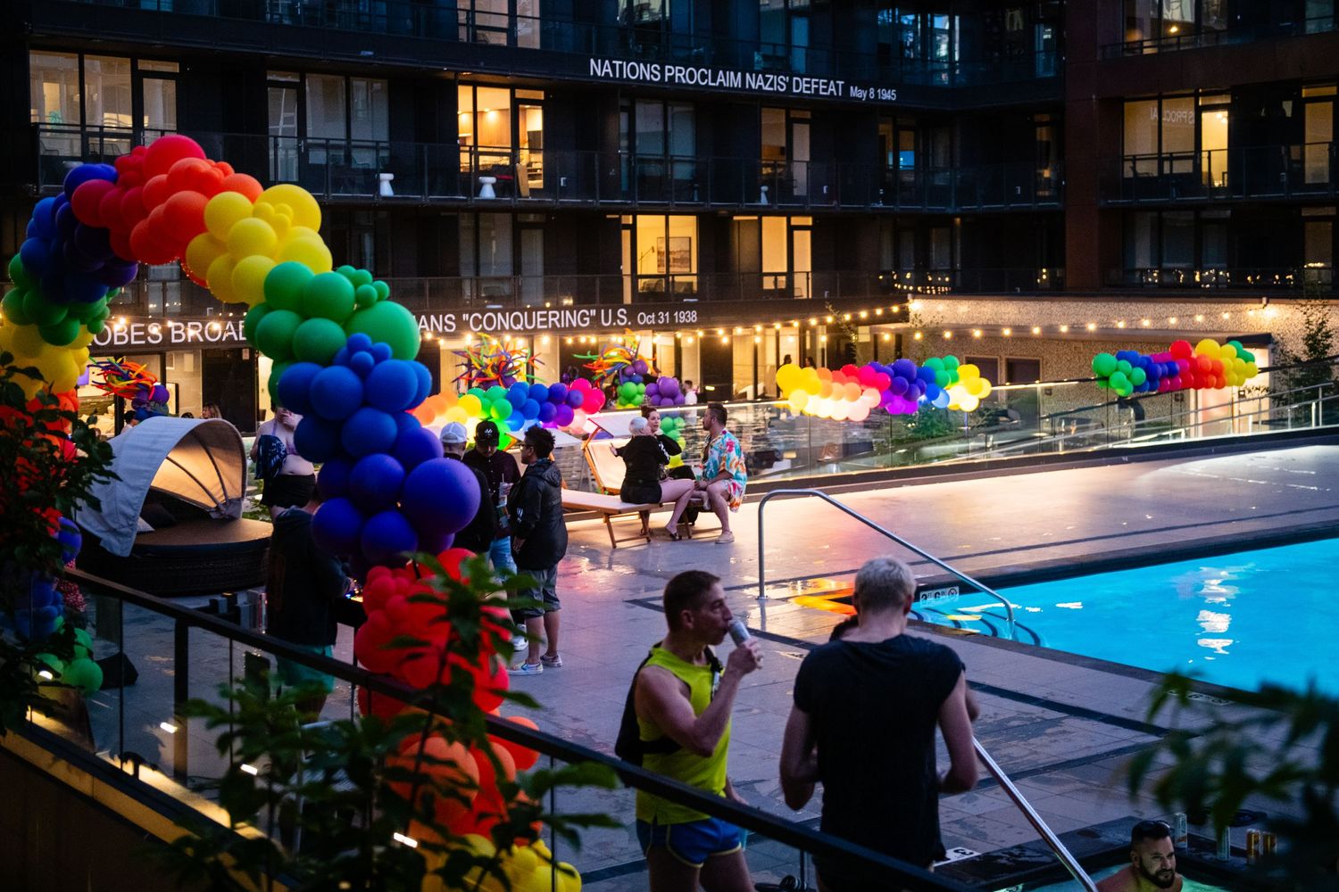 Colorful rainbow balloon arch decorates poolside evening party with glowing lights at urban apartment complex.