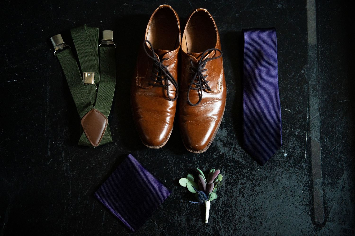 Wedding accessories including brown leather shoes, navy tie, and green suspenders arranged on dark surface.