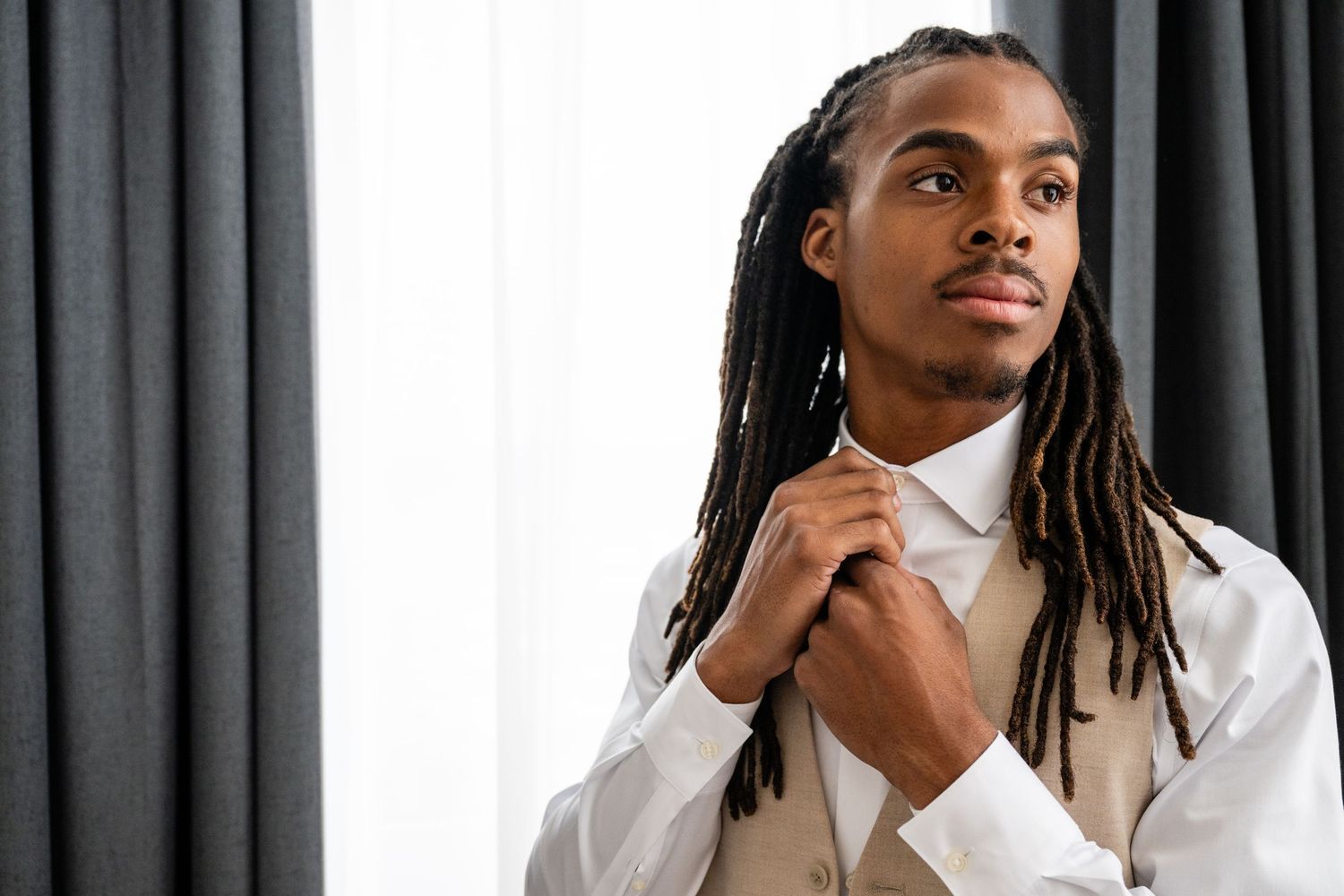 Professional portrait of someone in a white blazer with dreadlocks gazing thoughtfully against a gray backdrop.