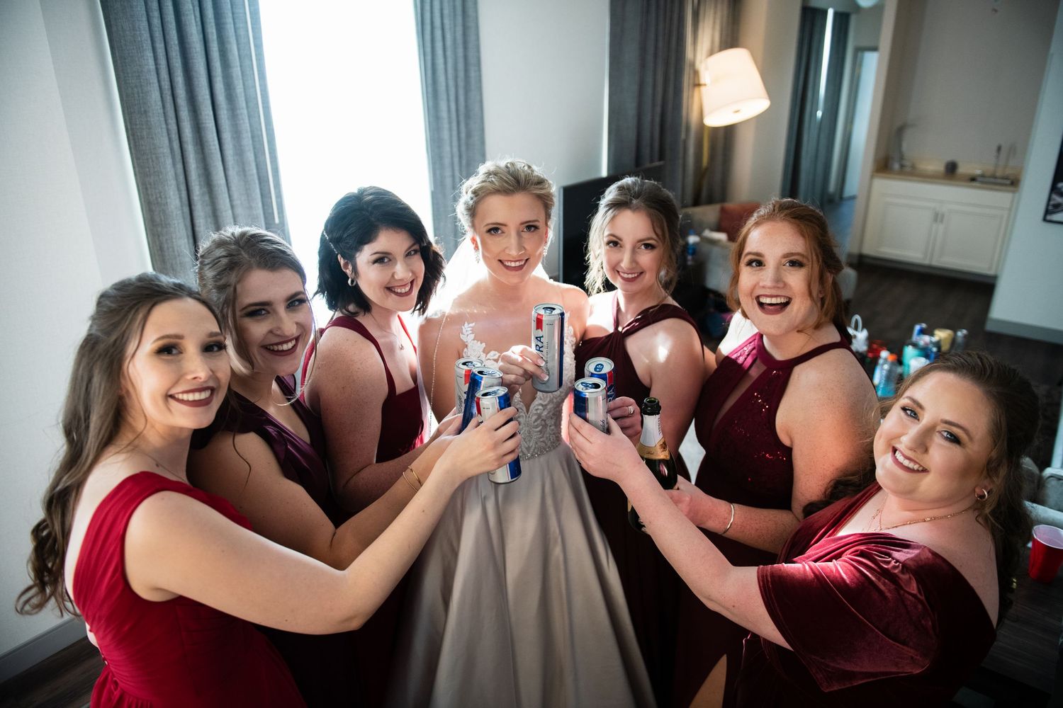 Wedding party toasting with drinks while surrounding the bride in burgundy bridesmaid dresses.