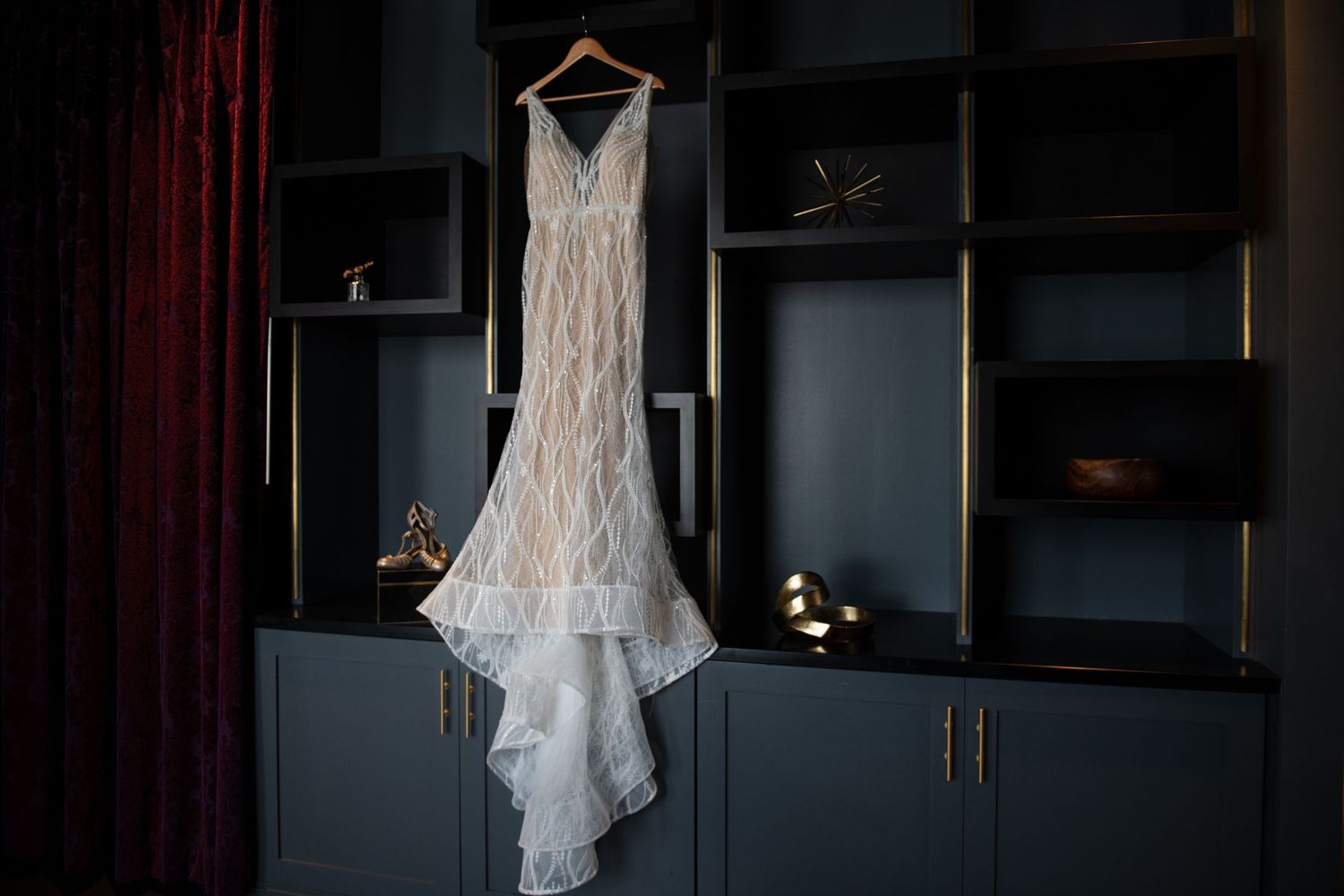 An elegant white lace wedding dress hangs against dark built-in shelving with dramatic lighting.