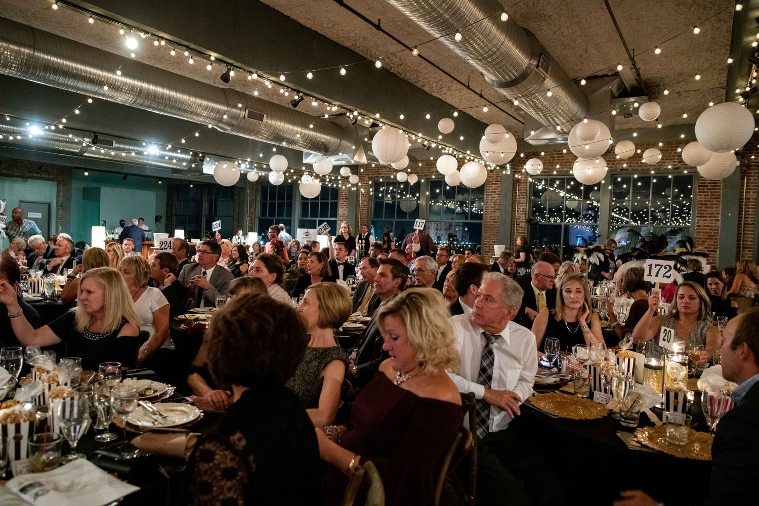 Large dining room filled with guests under paper lanterns and string lights for an elegant evening event.
