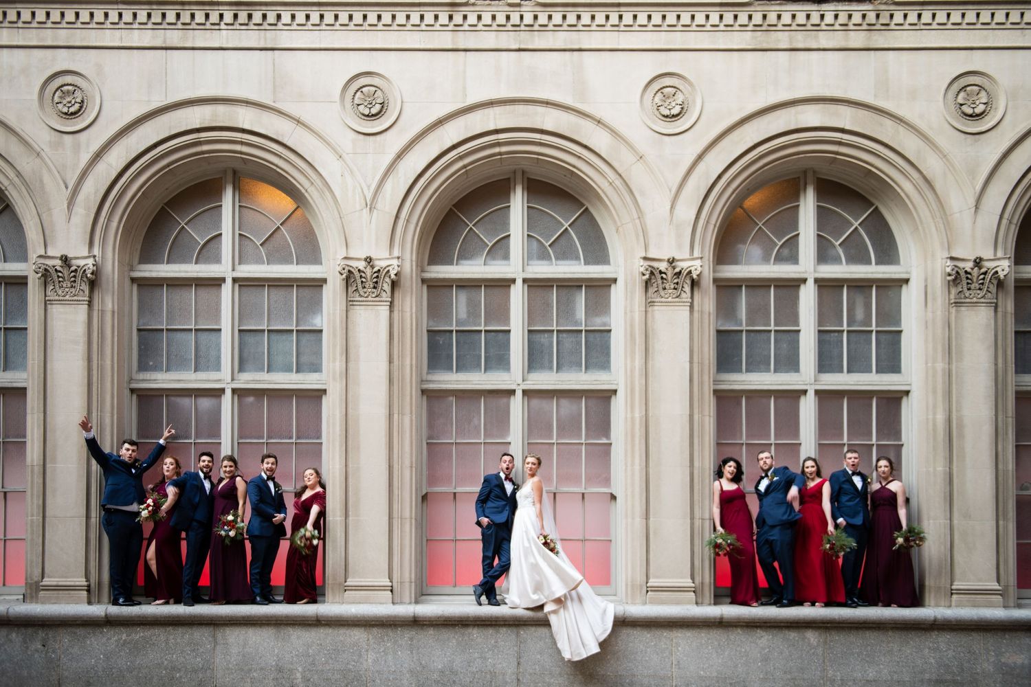 Wedding party poses creatively along arched windows of historic building facade.
