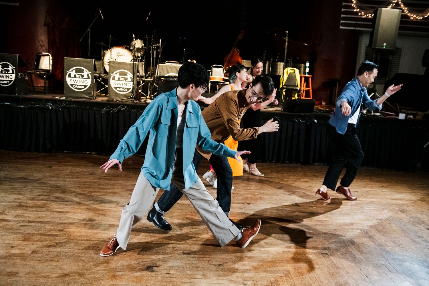 Dynamic dancers perform swing dance moves on a polished wooden floor in front of band equipment.