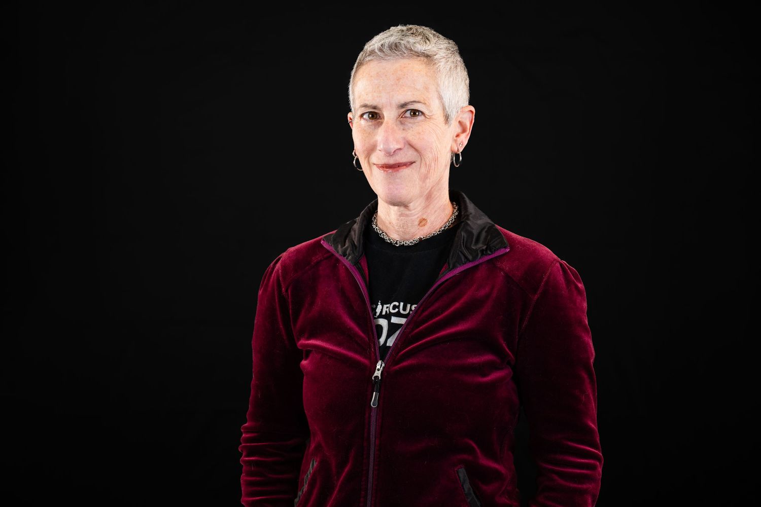 Professional portrait against black background showing person in burgundy velvet jacket and silver necklace.