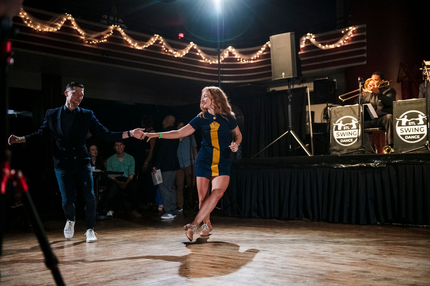 Dancers perform swing moves on wooden floor with string lights and band visible in background.