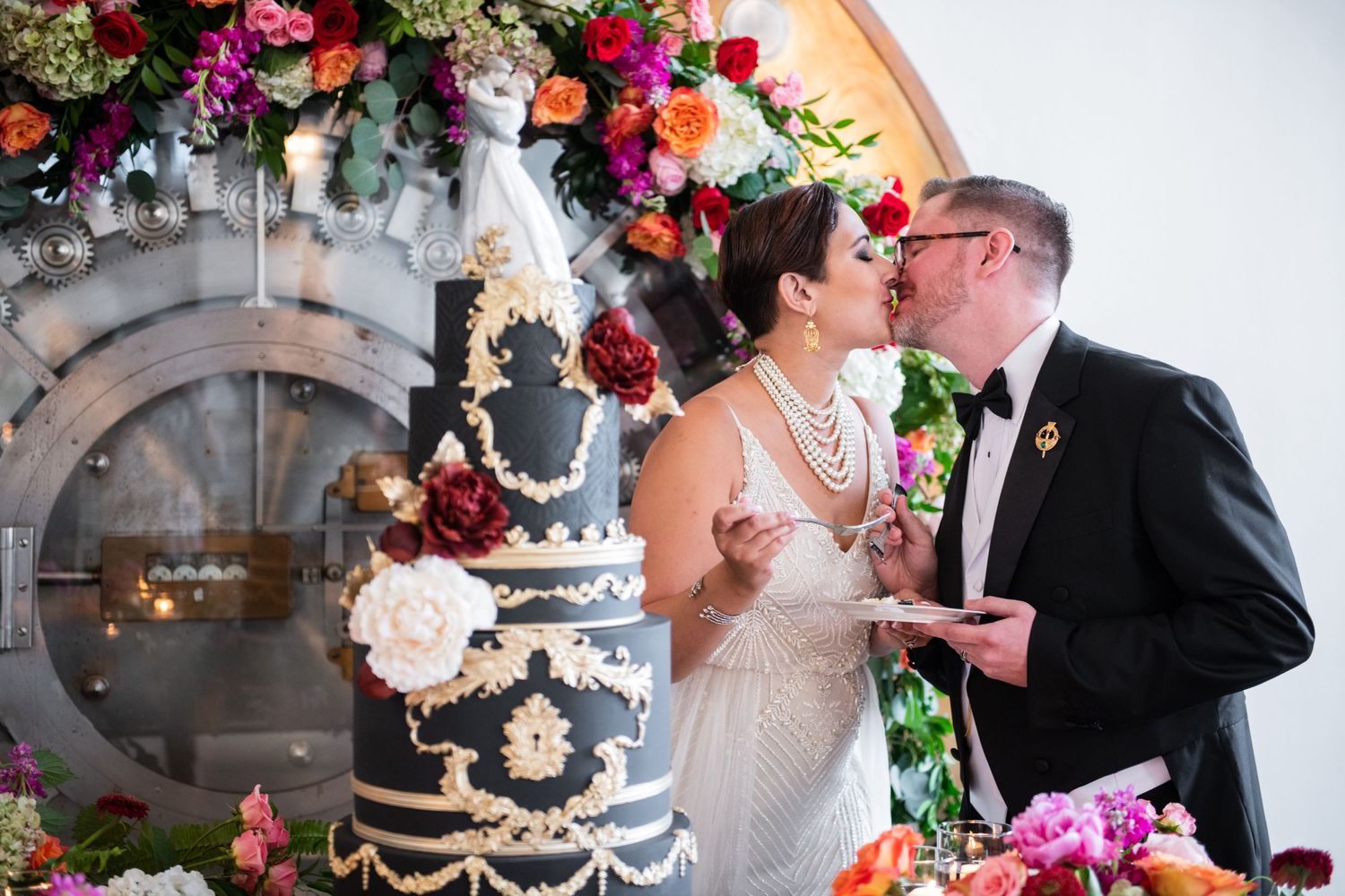 Wedding couple sharing a moment by their ornate black cake under vibrant floral arch decorations.