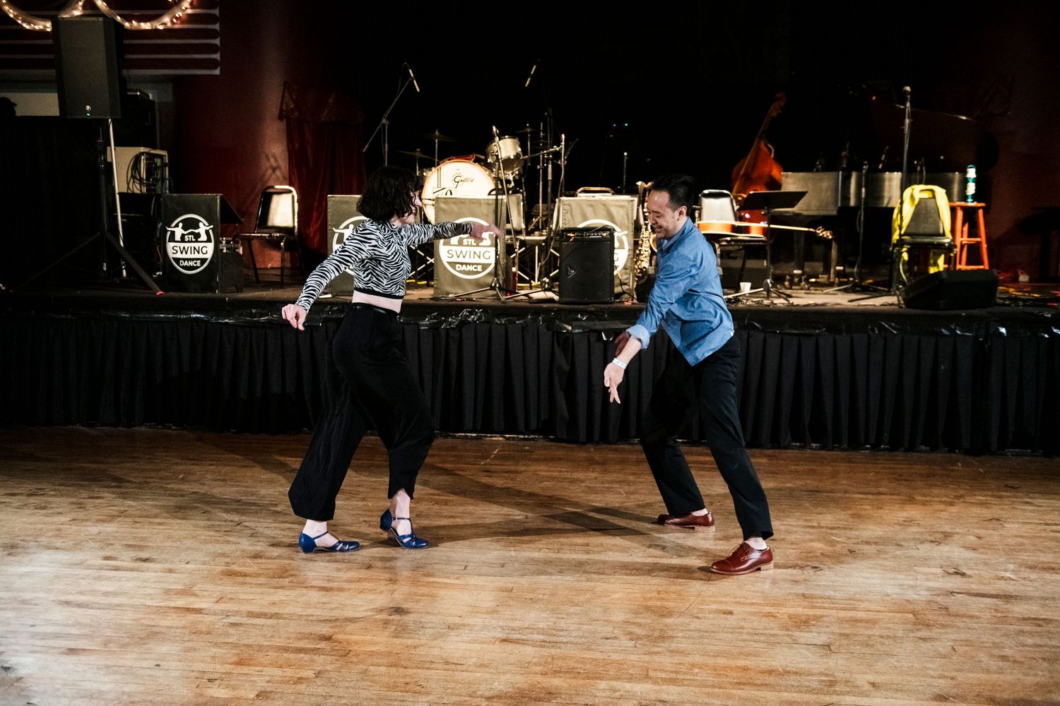 Two dancers perform swing dance moves on a wooden floor with stage equipment in the background.