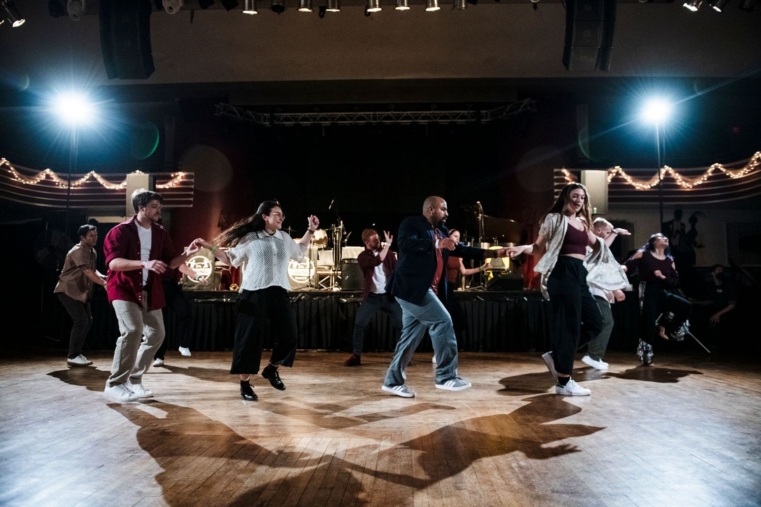 Dancers moving dynamically on a wooden dance floor with a live band performing in the background.