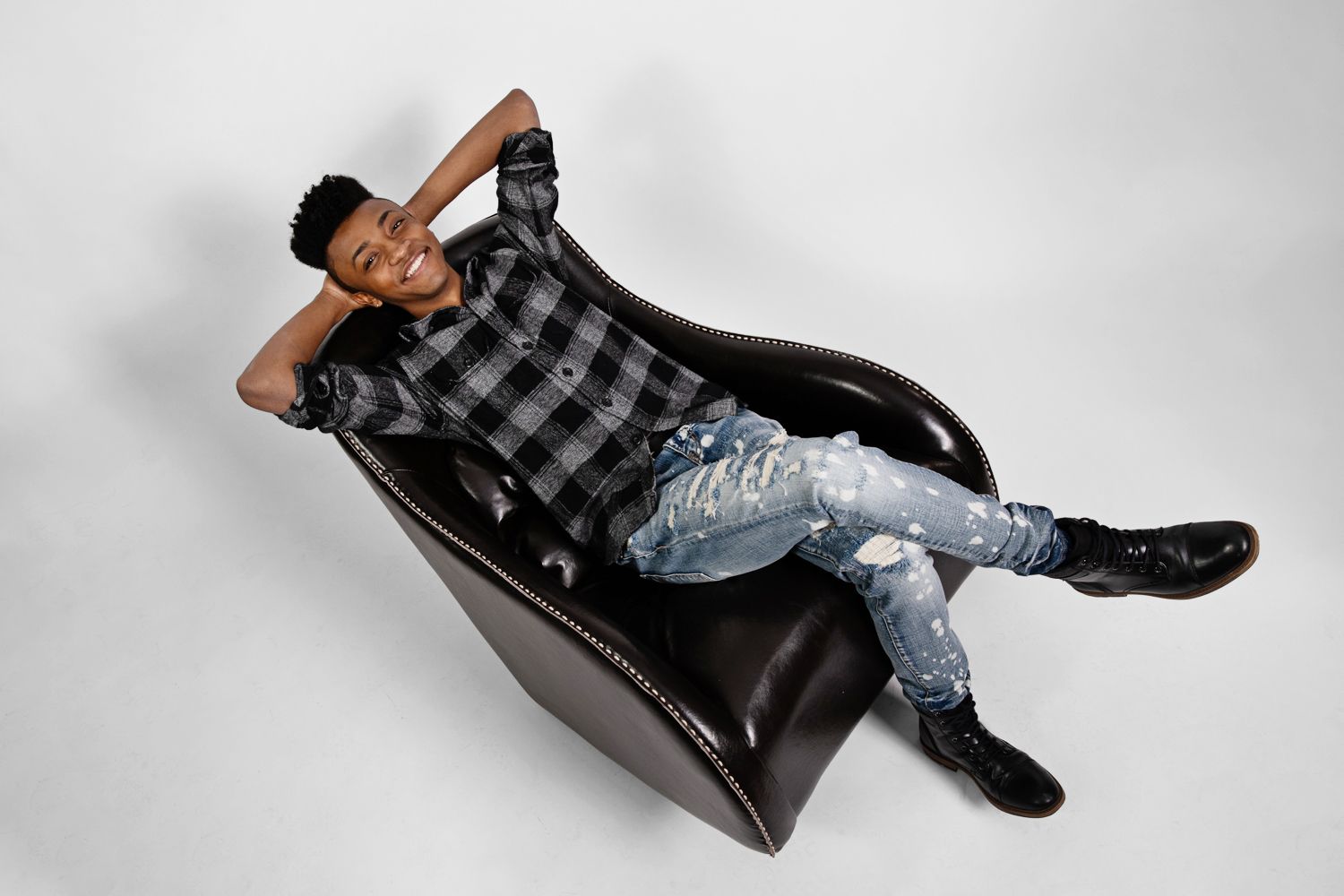 Person lounging in casual plaid shirt and ripped jeans on black leather chaise against grey background.