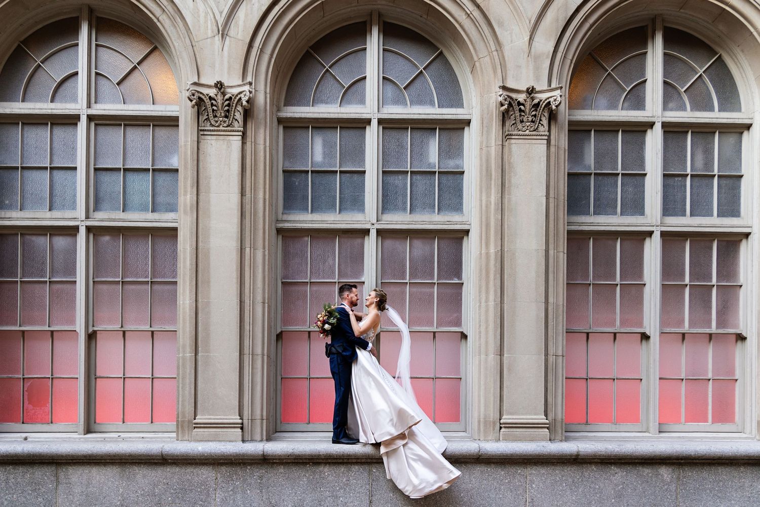 Romantic wedding couple poses against pink-lit arched windows in classic architecture.