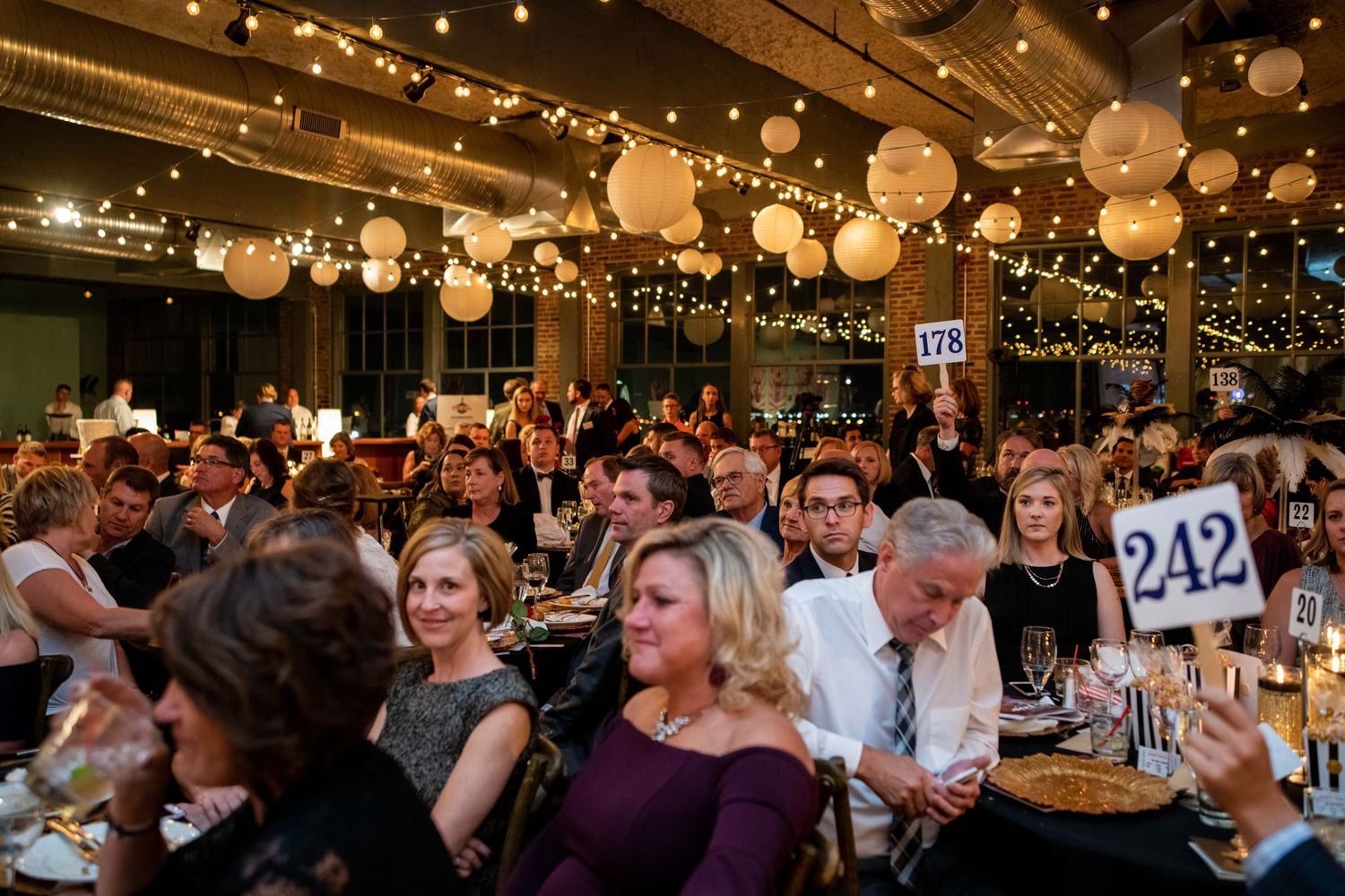Crowded formal dinner event with guests holding auction numbers under decorative paper lanterns.