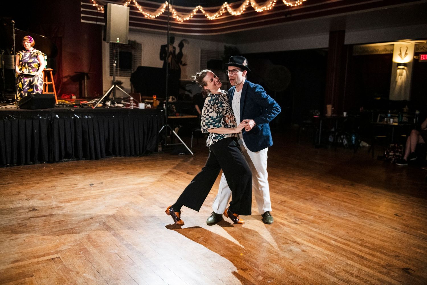 Dancers glide across wooden floor during swing dance performance with band in background.
