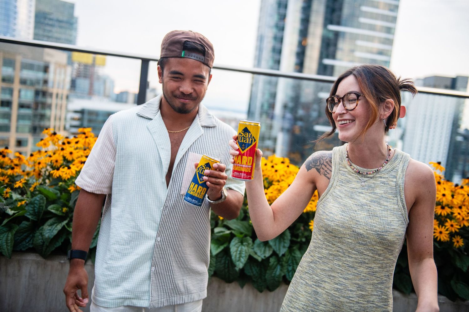 Two people toasting with beverages on an urban rooftop garden with sunflowers.