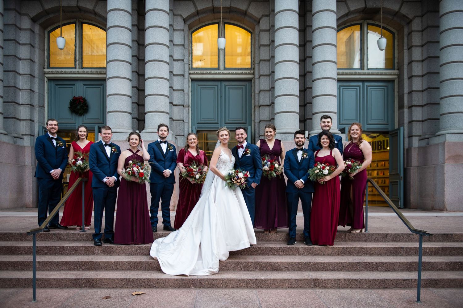 Wedding party in burgundy and navy poses on stone steps of historic building.