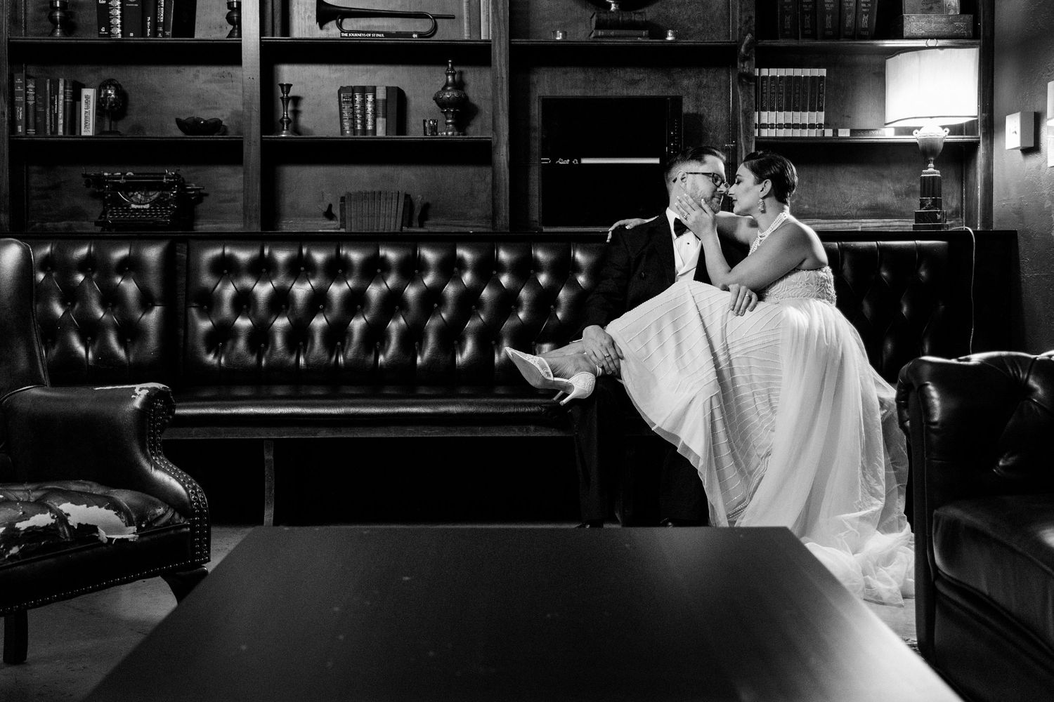 A black and white shot of a couple relaxing on a leather couch in a library setting.