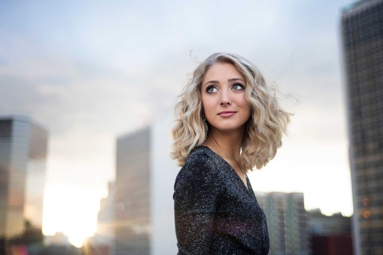 Person in sparkly black top looking to side with city skyline and sunset in background.