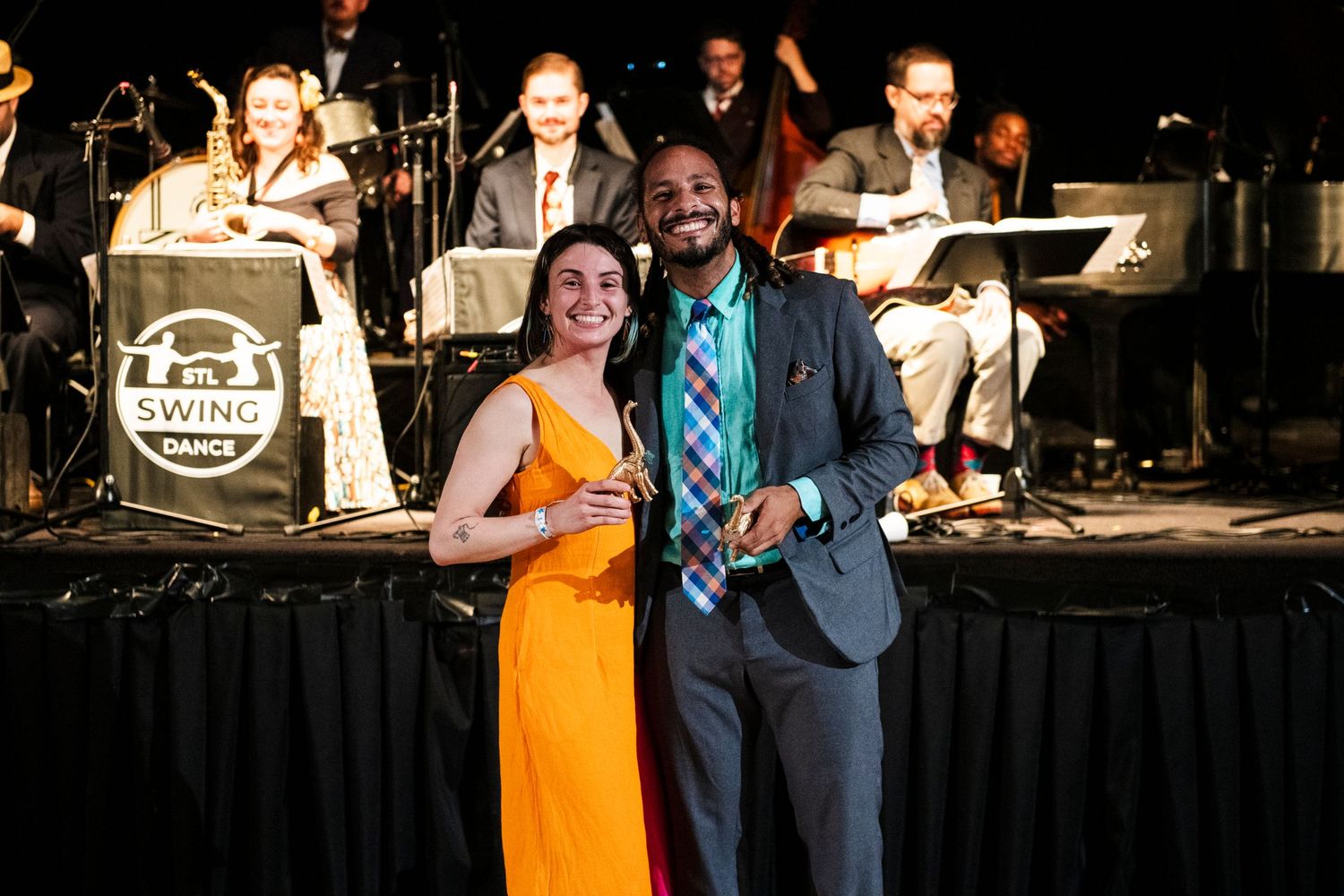 Performers pose in formal attire in front of big band orchestra on stage.