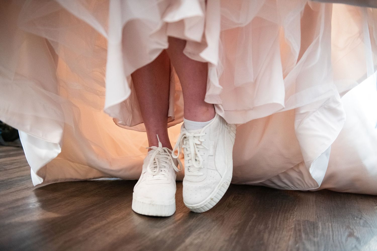 White sneakers peeking out under a flowing wedding dress on a hardwood floor.