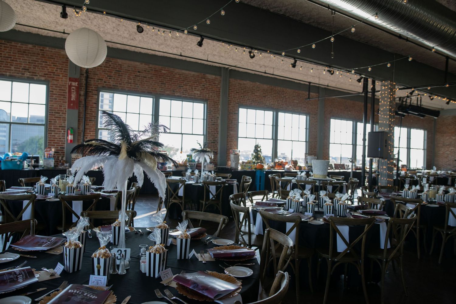 Industrial event space with palm tree decorations and elegant table settings near large windows.