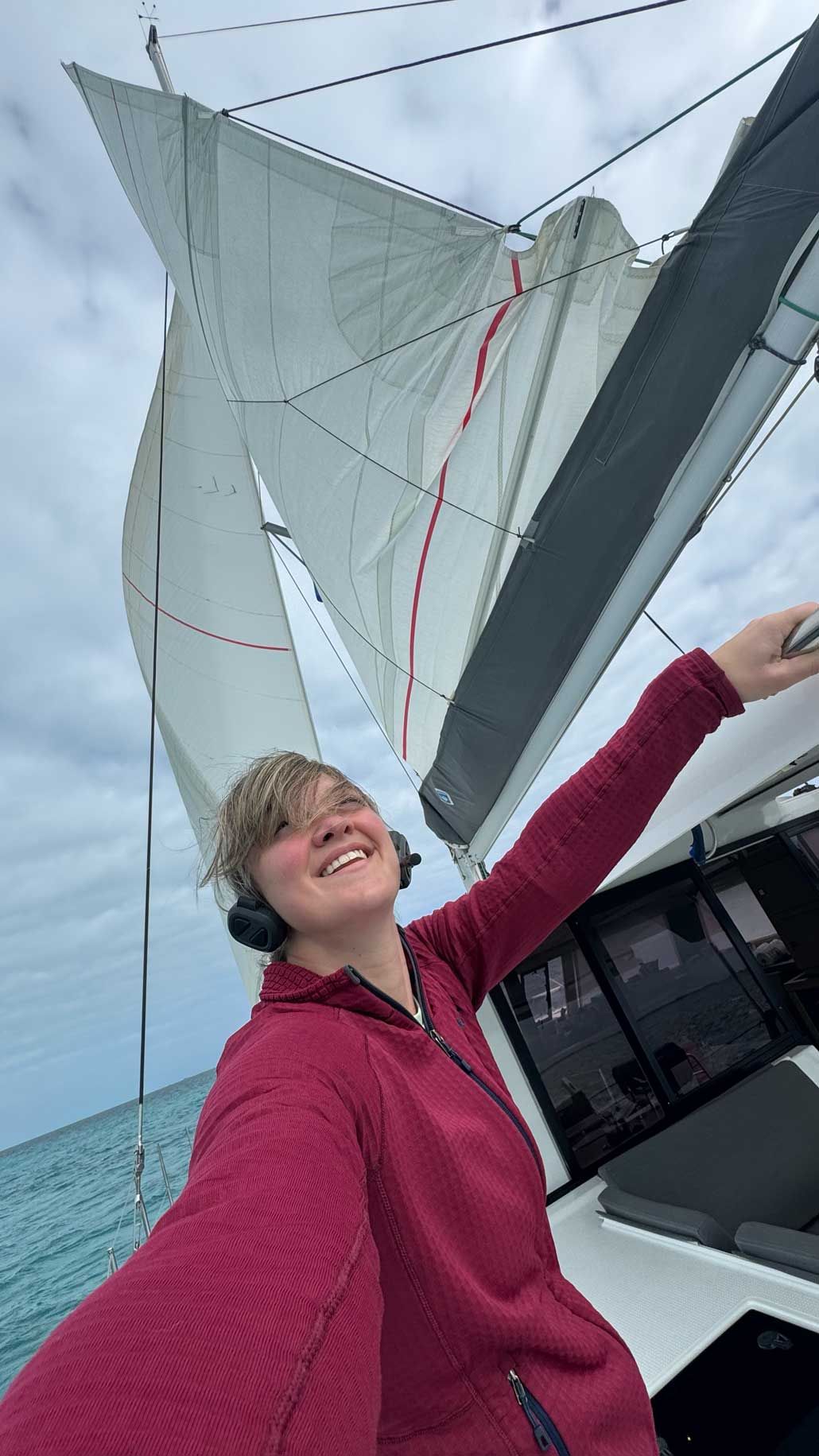 Smiling woman in red jacket on sailboat deck with white sails billowing against cloudy sky over turquoise ocean.