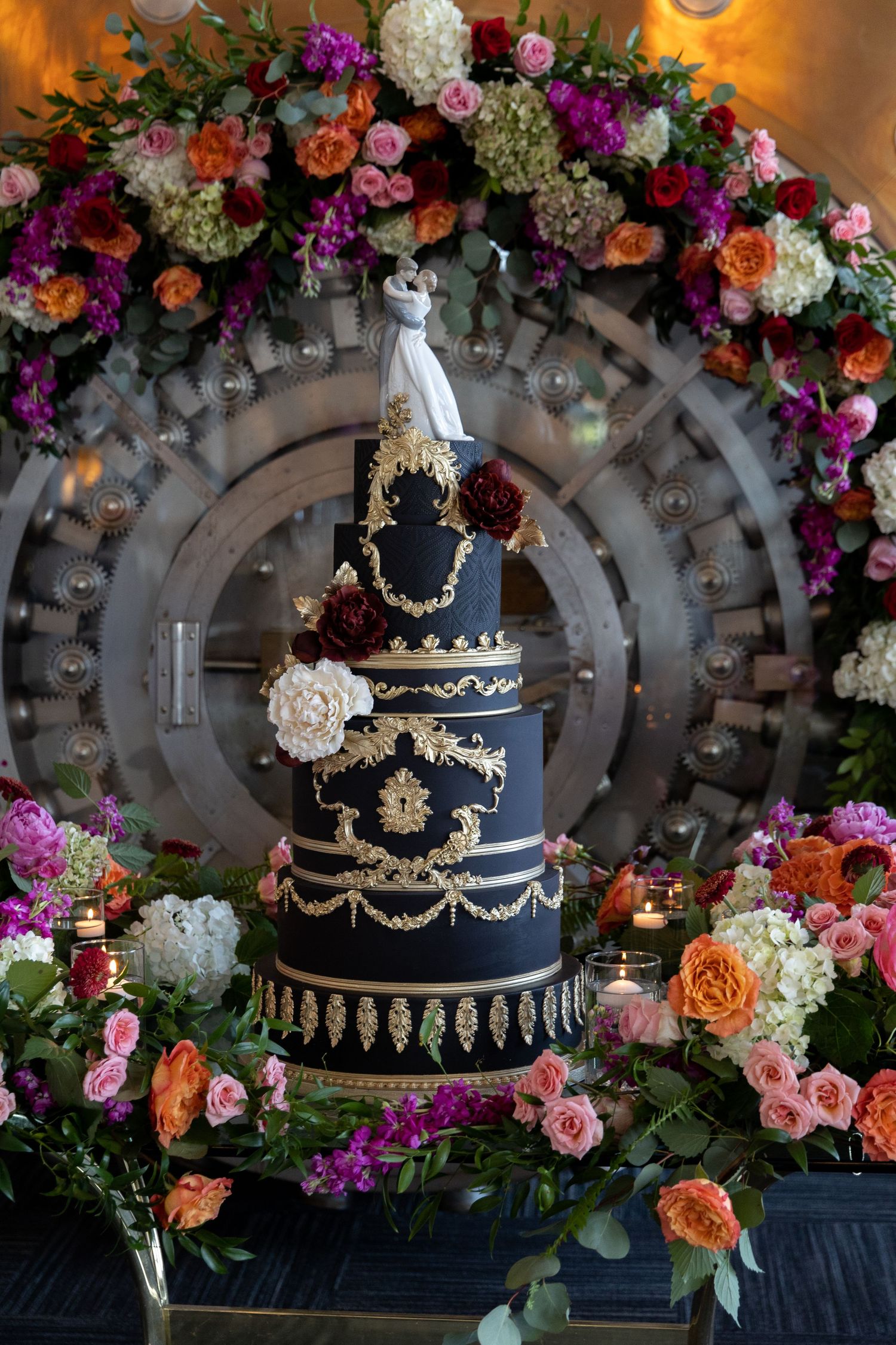 Elegant navy and gold wedding cake surrounded by vibrant floral arch display.