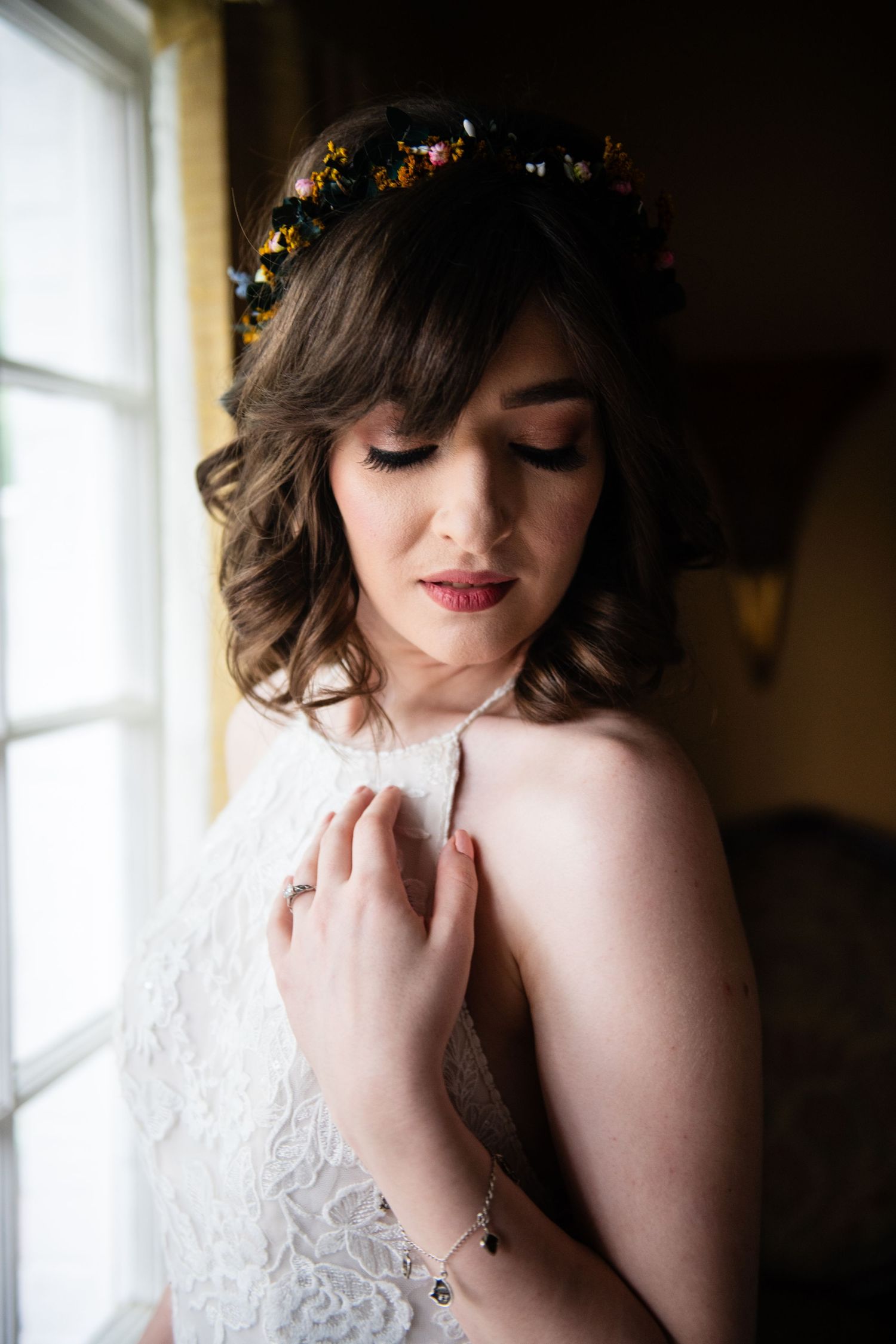 Bride in white lace dress with floral crown gazes down in romantic window portrait.