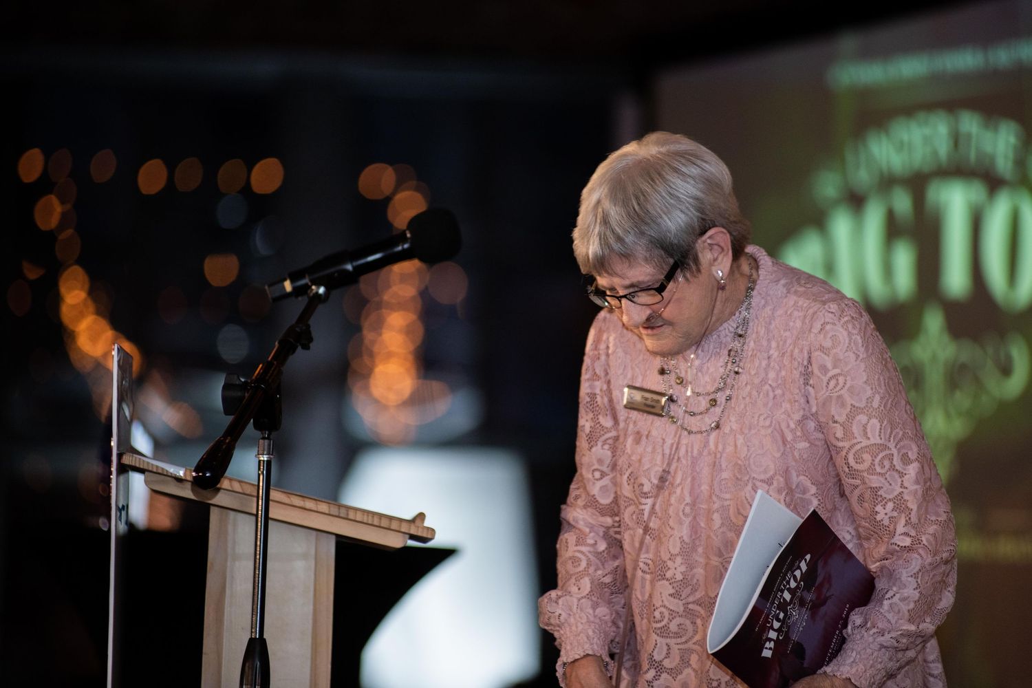 Speaker at wooden podium during evening event with bokeh lights in background.