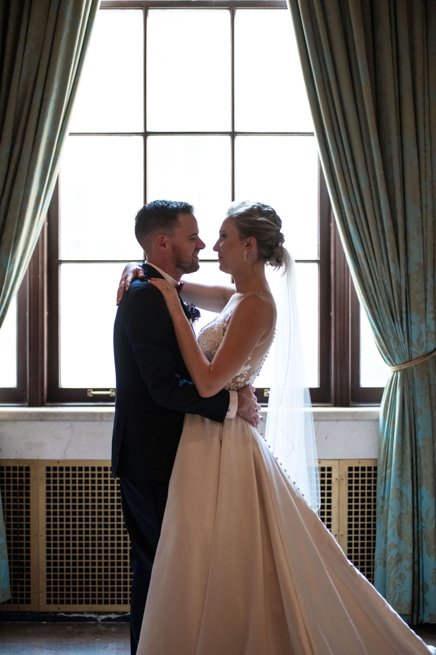 A romantic wedding couple embraces in front of a large window with elegant green curtains.