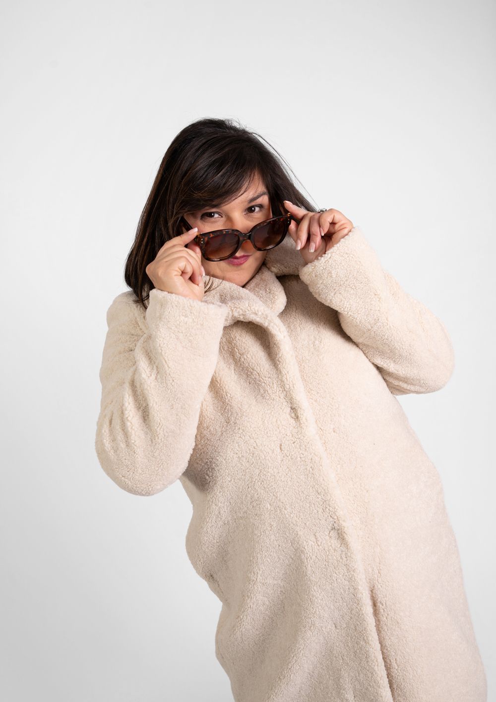 Woman in cozy cream coat playfully adjusting sunglasses against white background.