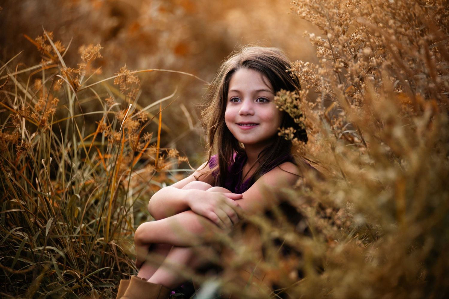 A portrait taken in warm autumn tones showing someone sitting among golden brown grasses and dried plants.