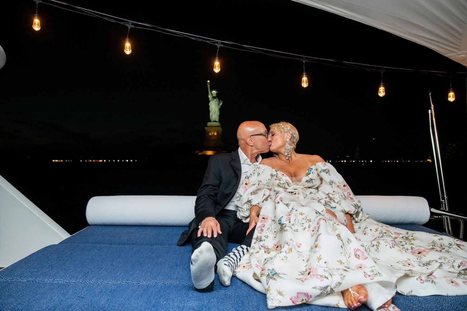 Couple relaxes on cushioned seating with Statue of Liberty visible in background at night.