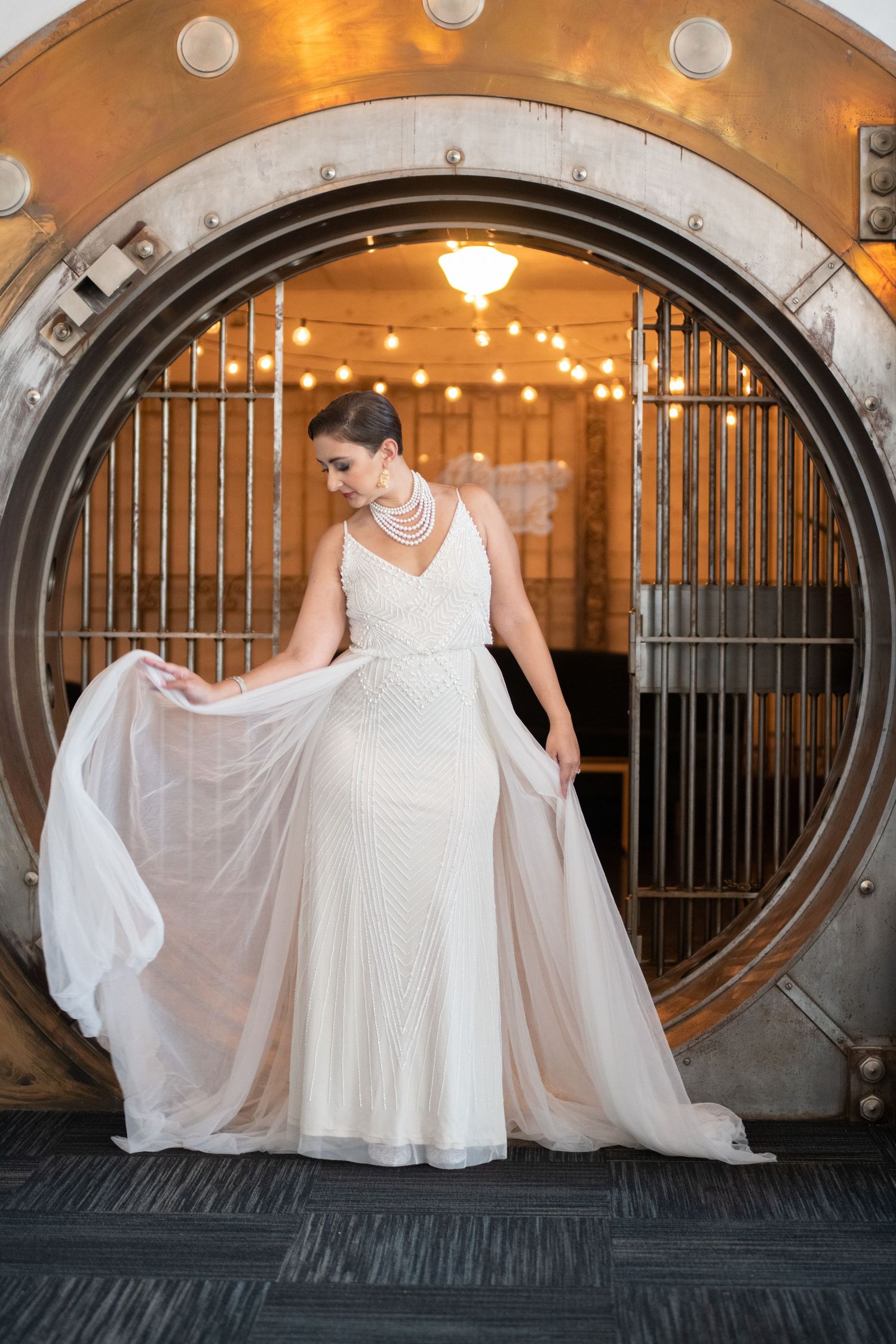 A bride in an elegant white dress poses in front of an illuminated vault door.