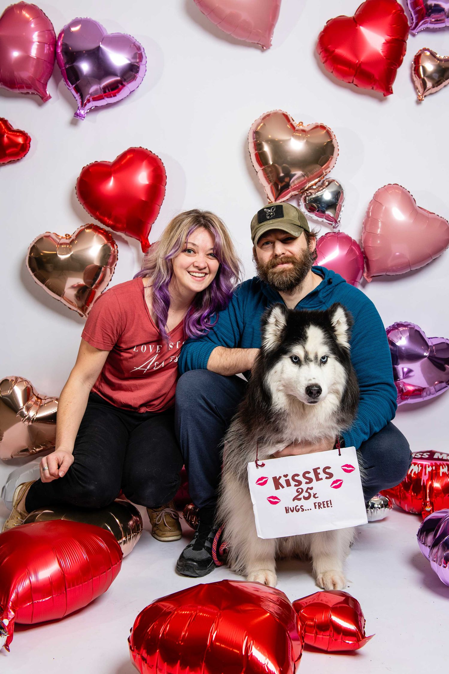 Husky dog poses with owners against backdrop of heart-shaped Valentine's Day balloons.