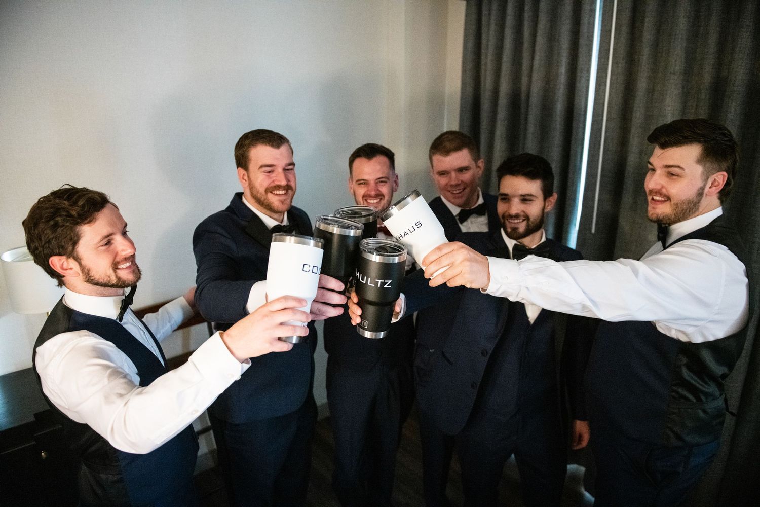 Groomsmen in matching vests toasting with drinks during wedding preparations.