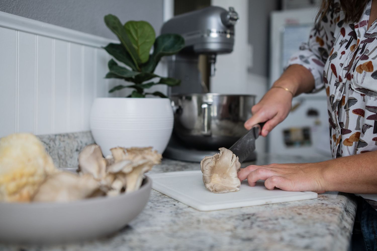 Hands chopping ingredients on a cutting board next to a marble countertop with a stand mixer.