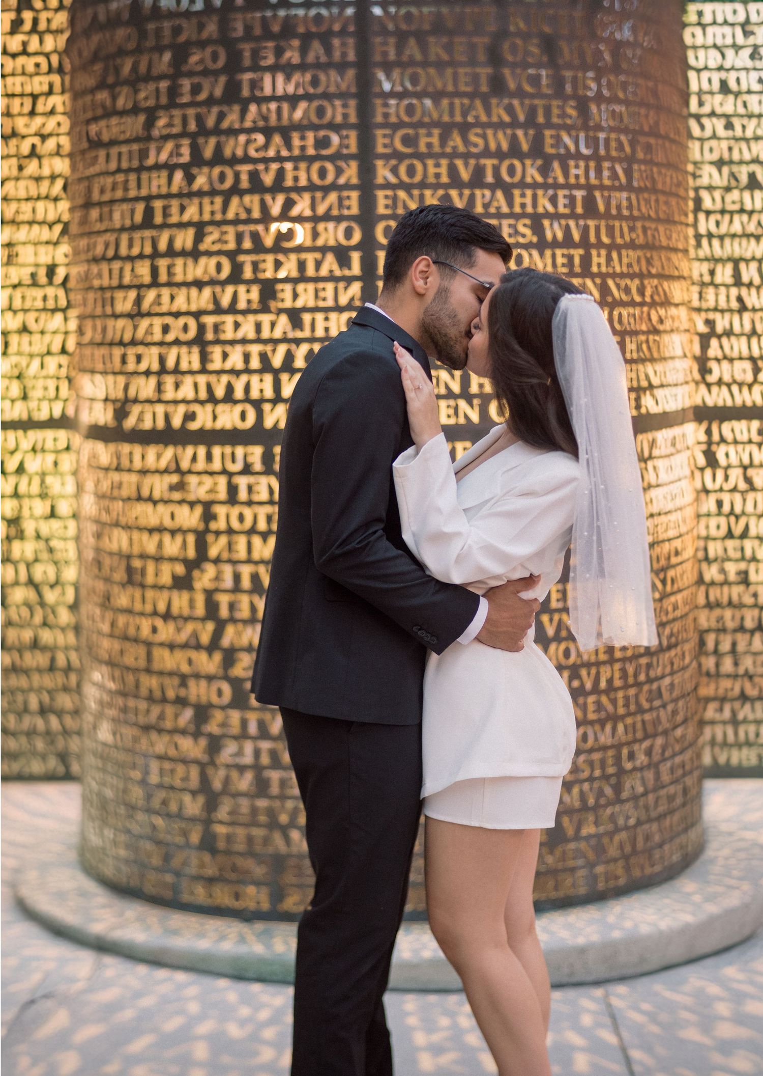 A romantic moment captured in front of a cylindrical monument with engraved text.
