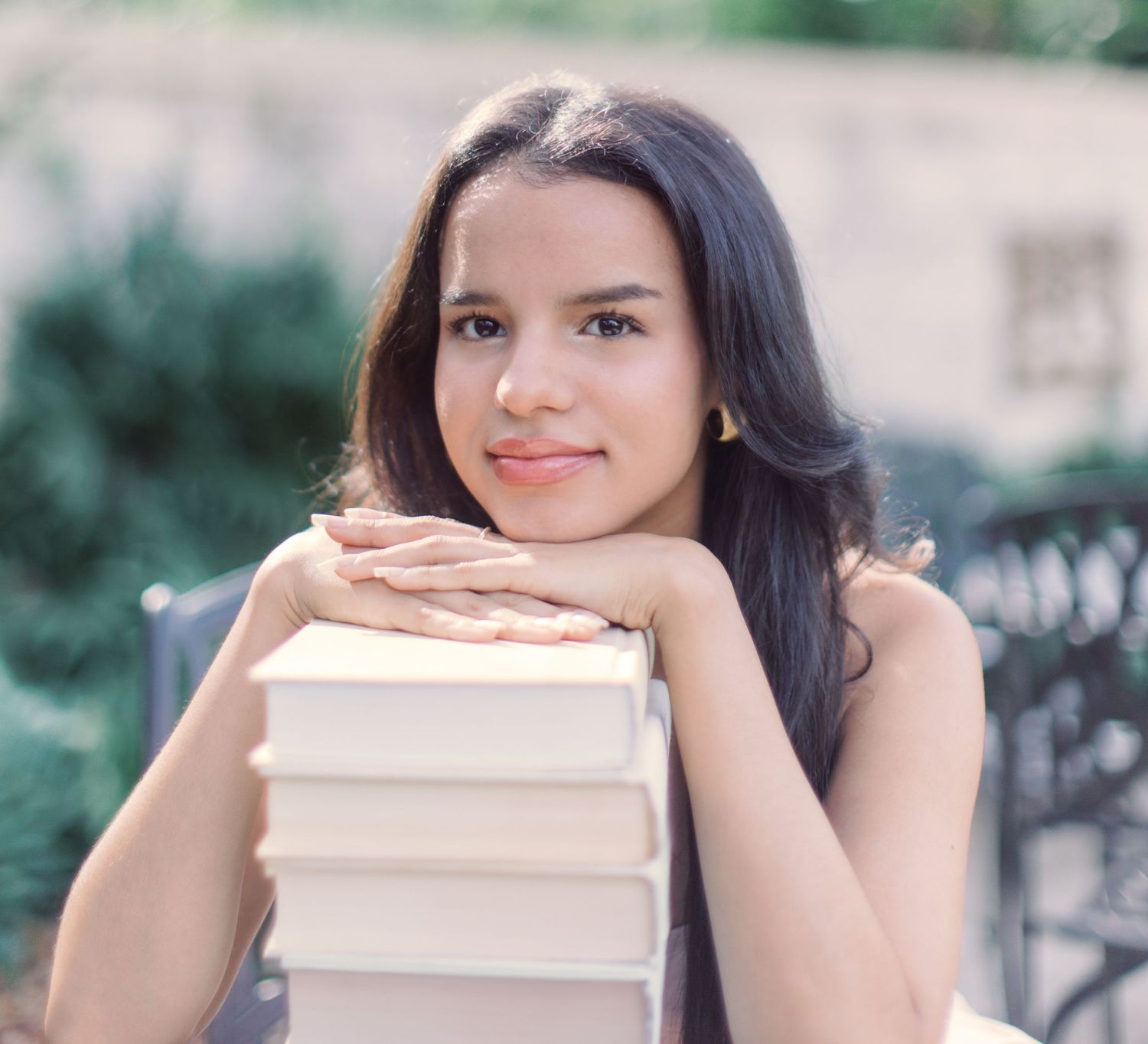 Long dark haired figure rests chin on stack of books with soft focus natural background.