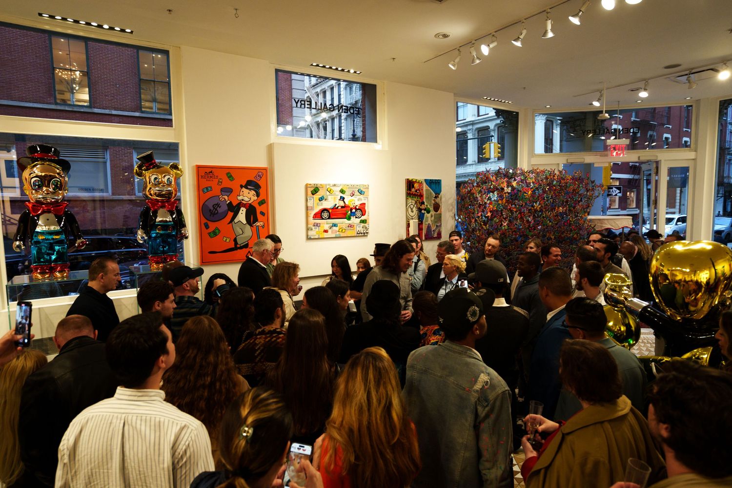 A crowded art gallery opening event with colorful paintings displayed on white walls and attendees mingling in the space.