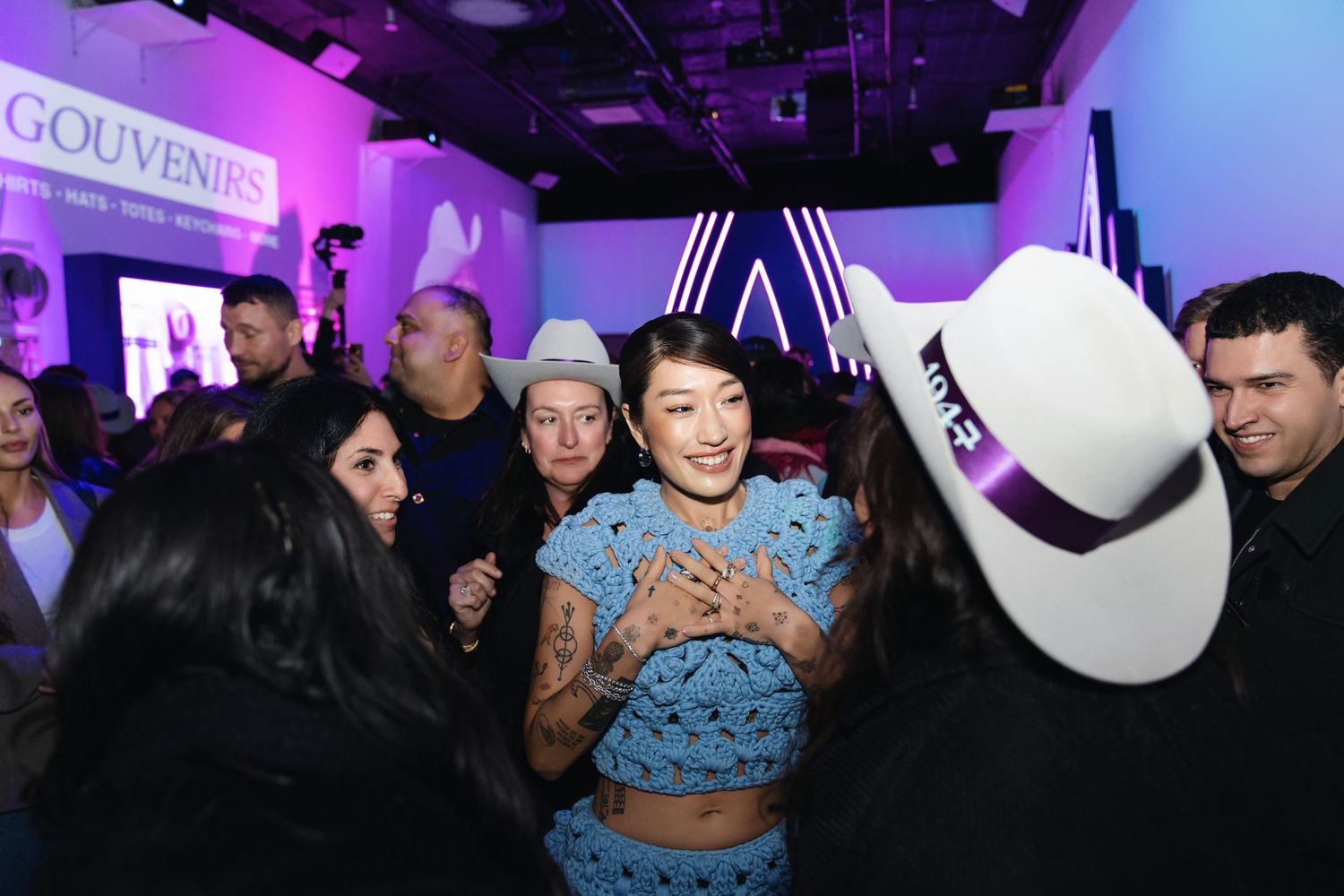 Crowded event scene with person in blue crochet outfit surrounded by guests in cowboy hats.