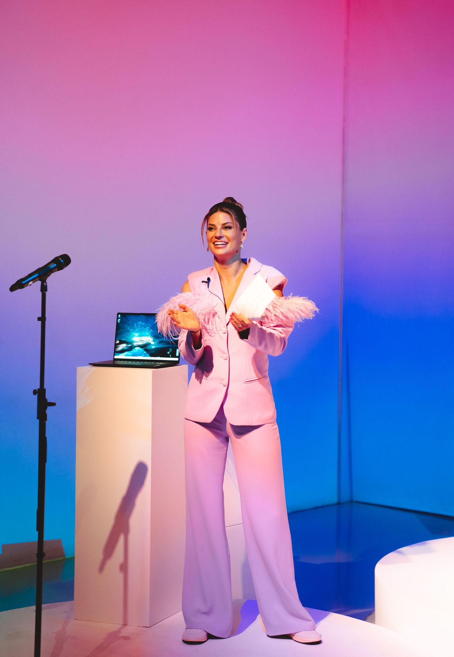 Presenter in pink suit speaks on stage with laptop display in pink and blue lit studio.