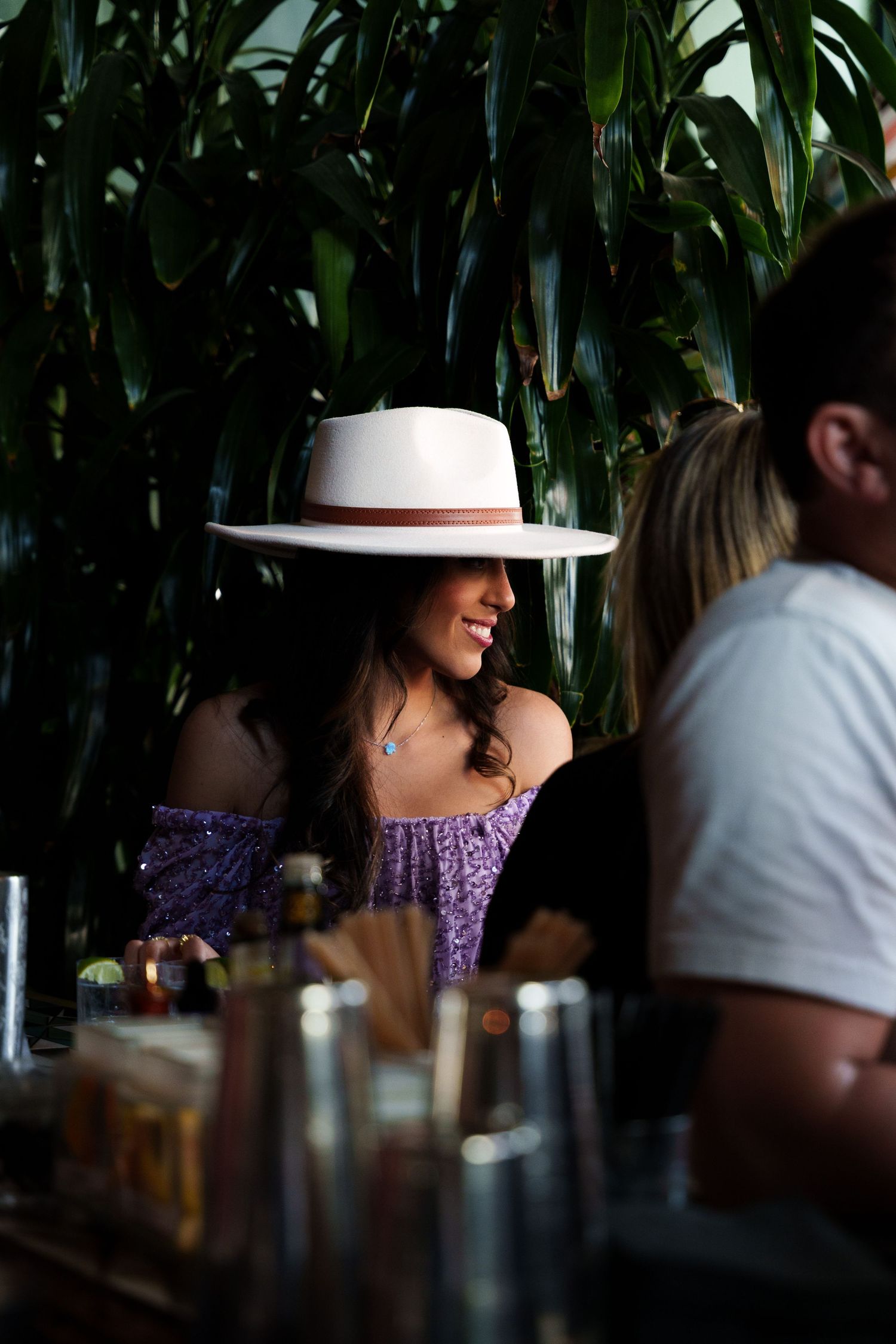 Person in purple dress and white hat seated at tropical-themed bar.