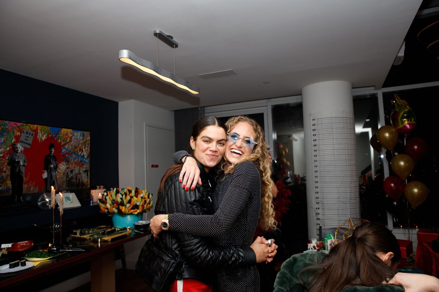 Two friends embrace at a party with festive decorations and lighting in the background.
