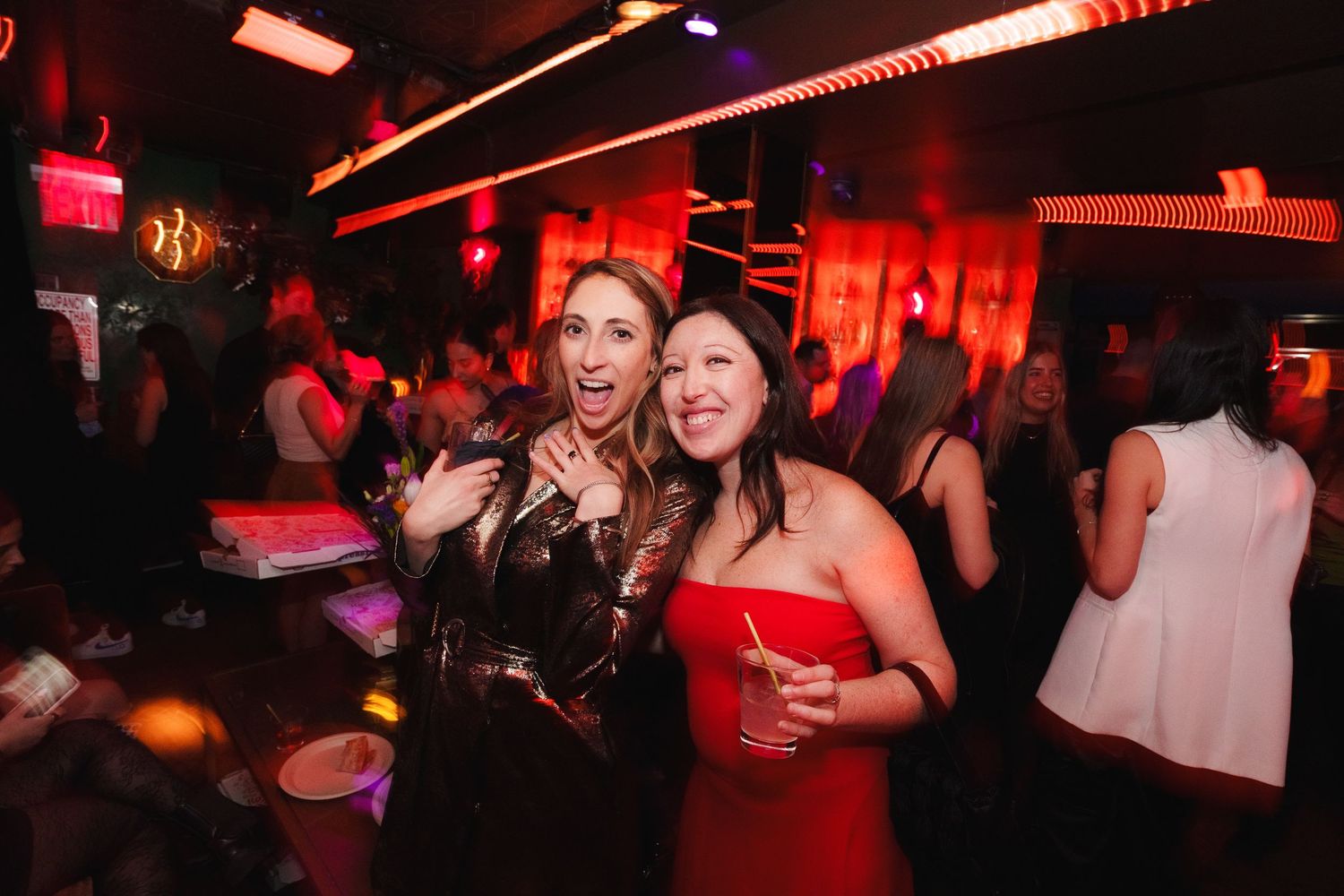 Partygoers celebrating at a nightclub with drinks under red and pink lighting.