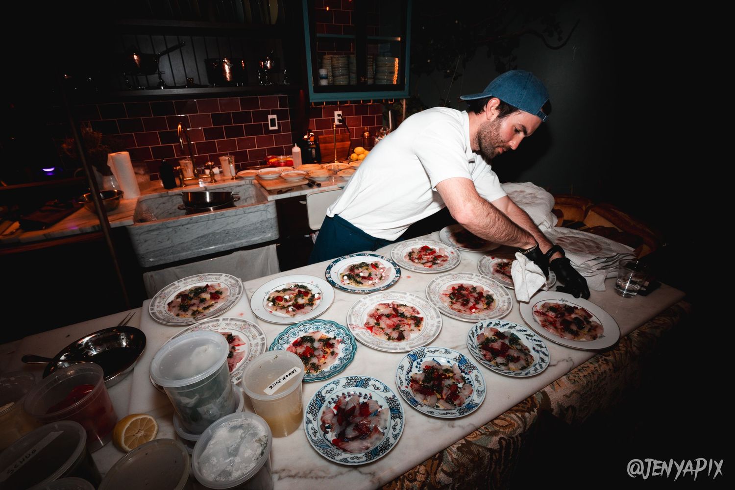 Chef plating multiple dishes on a dark counter in restaurant kitchen.