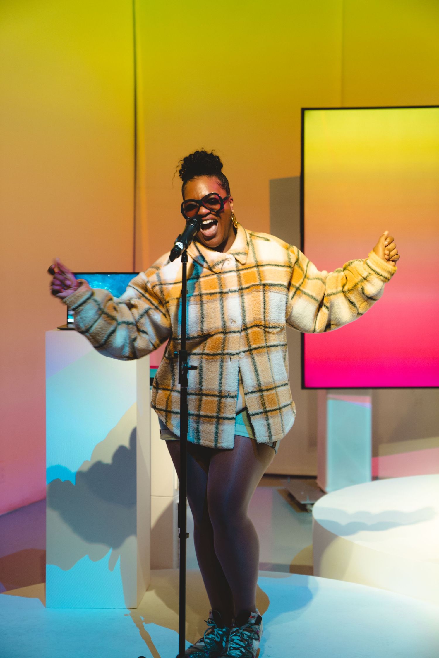 Performer in plaid shirt gestures enthusiastically on stage with colorful gradient backdrop.